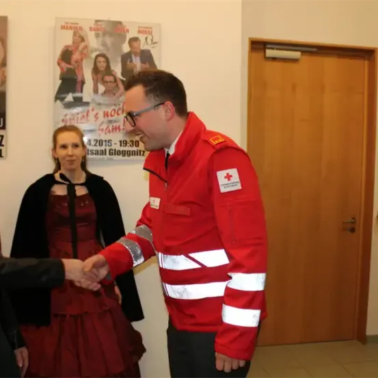 Ein Mann in einem roten Sicherheitsjackett schüttelt die Hand einer Frau in einem roten Kleid. Sie stehen vor einem Poster mit Datum und einer Tür.