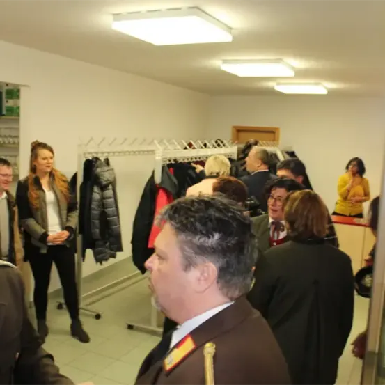 Eine Gruppe von Menschen versammelt sich in einem Raum mit weißen Wänden und Deckenleuchten. Einige sind in Uniformen, andere in Freizeitkleidung. Sie betrachten Kleidung, die an einer Garderobe hängt.