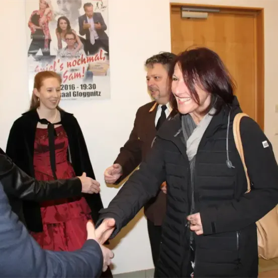 Fünf Menschen stehen in einem Raum und schütteln Hände. Eine Frau in Schwarz schüttelt die Hand eines Mannes in Anzug. Eine andere Frau trägt ein rotes Kleid. Ein Poster ist an der Wand.