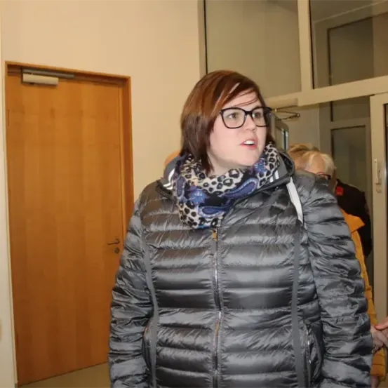 Eine Frau mit Brille und Schal läuft in einem Gebäude, möglicherweise in einem Flur. Sie hat einen ernsten Gesichtsausdruck. Hinter ihr steht eine Person in der Nähe einer Tür.