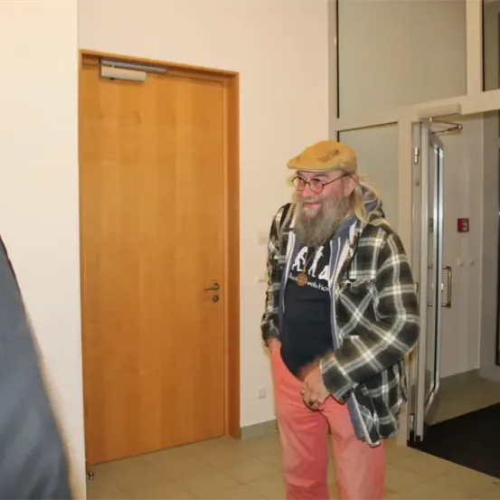 Ein bärtiger Mann mit Hut steht lächelnd vor einer Tür.