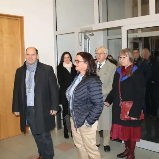 Eine Gruppe Erwachsener, in Winterkleidung gekleidet, versammelt sich in einem Innenraum, möglicherweise bei einer gesellschaftlichen Veranstaltung. Sie stehen in der Nähe einer offenen Tür.