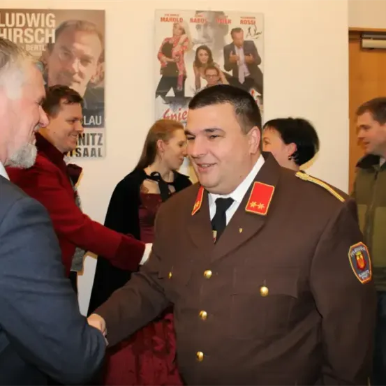 Eine Gruppe von Menschen ist drinnen. Ein Mann in Uniform schüttelt einem Mann im Anzug die Hand. Hinter ihnen stehen mehrere Menschen, einige in formeller Kleidung. Poster hängen an der Wand.