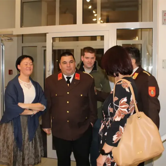 Fünf Menschen stehen vor einer Tür. Der Mann in der Mitte trägt eine Militäruniform. Zwei Frauen stehen auf der linken und rechten Seite. Eine Frau auf der rechten Seite trägt eine Handtasche. Die Tür hat ein Schild, das 'ZUGAN' besagt.