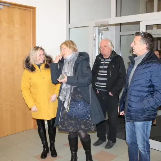 Vier Menschen stehen in einem Flur, zwei Frauen und zwei Männer. Sie scheinen ein Gespräch zu führen. Die Frau auf der linken Seite lächelt, während die andere Frau ein Handy in der Hand hält. Die Männer schauen in die gleiche Richtung.