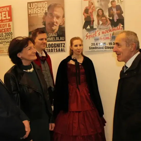 Eine Gruppe von Menschen steht vor einer Wand mit verschiedenen Postern, eines davon zeigt Ludwig Hirsch.