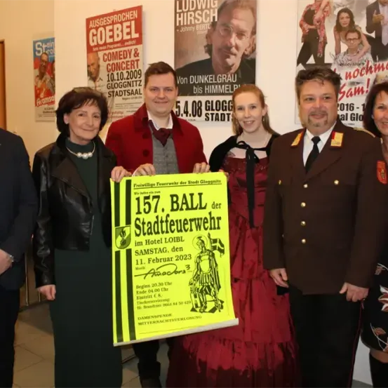 Eine Gruppe Erwachsener steht vor einer Wand mit Postern und hält ein gelbes Schild für den 157. Ball der Stadtfeuerwehr am 11. Februar 2023 im Hotel LOIB.