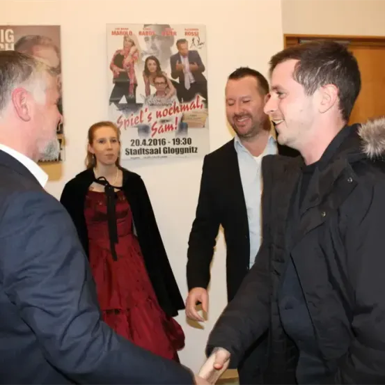 Vier Personen stehen vor einer Wand, lächeln und schauen sich gegenseitig an. Der Mann auf der rechten Seite gibt dem Mann auf der linken Seite die Hand. Es gibt zwei Poster an der Wand. Eines davon hat ein Bild von einem Mann und einer Frau, und das andere hat einen Text in einer fremden Sprache.