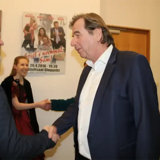 Zwei Männer schütteln sich die Hände vor einem Plakat. Der Mann auf der rechten Seite trägt einen dunklen Anzug, während der Mann auf der linken Seite einen schwarzen Anzug trägt. Hinter ihnen steht eine Frau in einem roten Kleid in der Nähe einer anderen Person.