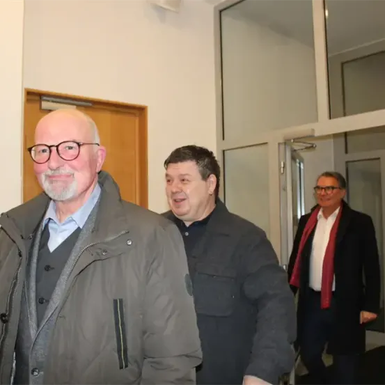 Drei Männer stehen in einem Flur. Der Mann in der Mitte trägt einen grauen Mantel. Der Mann auf der rechten Seite trägt einen schwarzen Mantel und einen roten Schal.