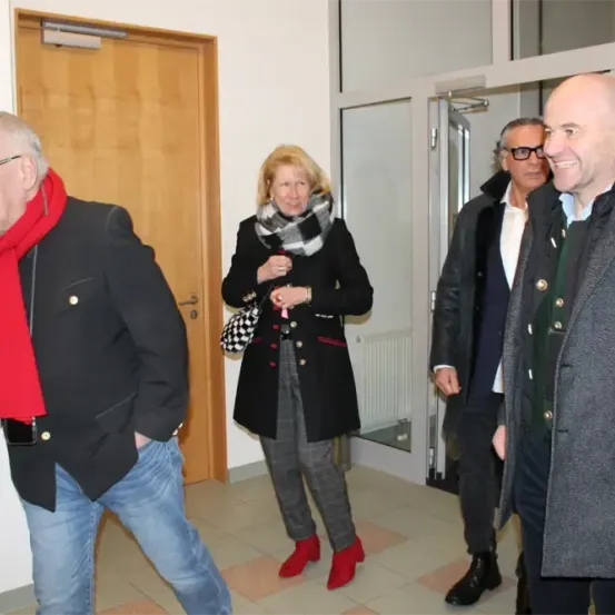 Vier Personen in Winterkleidung stehen in einem Flur und führen vermutlich ein Gespräch. Die Frau in der Mitte trägt einen Schal und eine Handtasche. Der Mann rechts lächelt und trägt eine Brille.