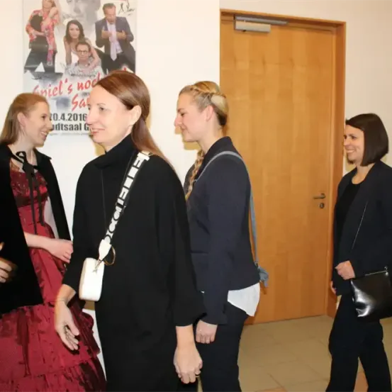 Vier Frauen unterhalten sich in der Nähe einer Tür, eine in einem roten Kleid und drei in Schwarz. Ein Poster hinter ihnen zeigt eine Gruppe von Menschen.