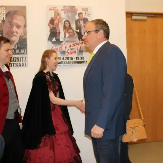 Vier Personen in einem Raum, zwei Erwachsene schütteln sich die Hände, während die anderen beiden lächeln. Ein Poster mit einem Bild eines Mannes und einer Frau ist an der Wand.