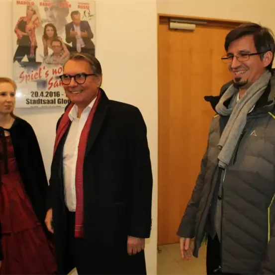 Drei Personen stehen vor einem Plakat für ein Theaterstück, zwei Männer lächeln und eine Frau schaut sie an.