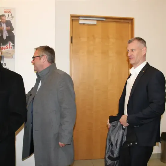 Drei Männer stehen in einem Flur und unterhalten sich. Hinter ihnen befindet sich ein Poster eines Mannes mit einem Schild, das 'mal' steht. Ein Mülleimer befindet sich rechts.