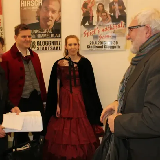Eine Gruppe von Menschen steht vor einer Wand mit Plakaten. Eine Person hält ein Papier und lächelt, eine andere trägt ein rotes Kleid und einen schwarzen Umhang.