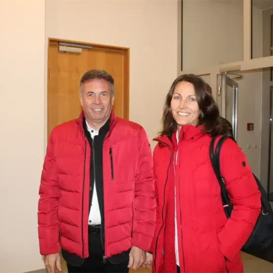 Zwei Personen, ein Mann und eine Frau, stehen eng beieinander in einem Flur, beide tragen rote Jacken und lächeln für die Kamera.