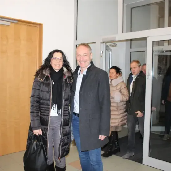 Ein Mann und eine Frau lächeln für ein Foto in einem Flur, mit Menschen, die im Hintergrund gehen.