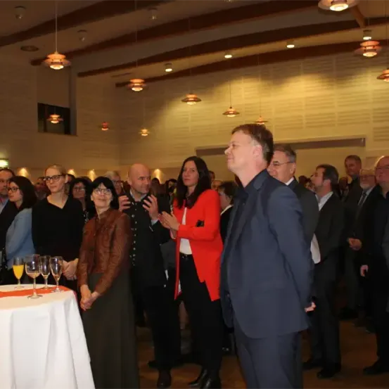 Eine formelle Veranstaltung mit mehreren Personen, die stehen, klatschen und eine Person nach oben schaut. Es gibt Gläser mit Orangensaft auf einem Tisch, und die Leute tragen formelle Kleidung.