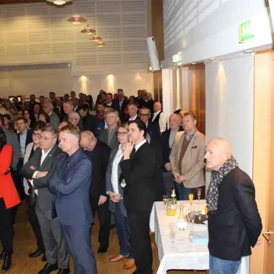 Eine große Gruppe formell gekleideter Menschen steht in einem Raum. Einige unterhalten sich, während andere zusehen. Ein Tisch mit Gläsern und Flaschen steht vor einer geschlossenen Tür.