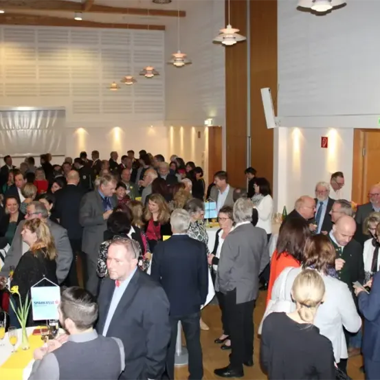 Eine große Gruppe von Menschen in formeller Kleidung, wahrscheinlich bei einer Konferenz, unterhalten sich und bewegen sich. Der Raum ist gut beleuchtet, und Tische mit gelben Tischdecken sind aufgestellt. Das Bild fängt eine dynamische und lebhafte soziale Atmosphäre ein.