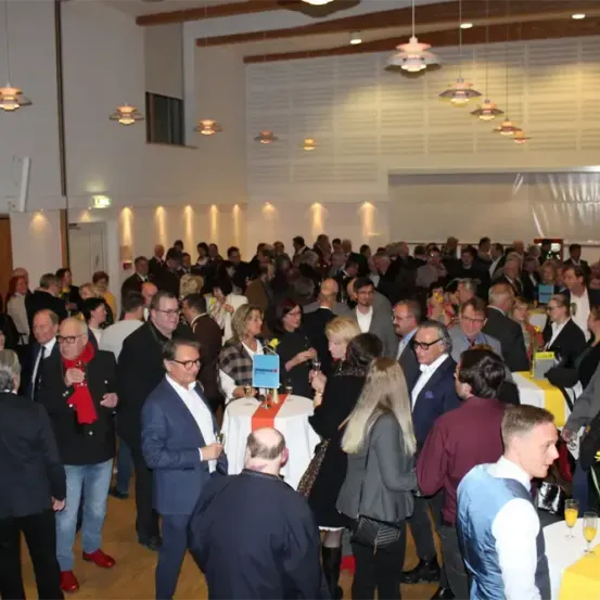 Eine große Menschenmenge formell gekleideter Männer und Frauen mischt sich in einem gut beleuchteten Innensaal mit weißen Wänden und Holzbalken, gelb gedeckten Tischen und hängenden Lampen.