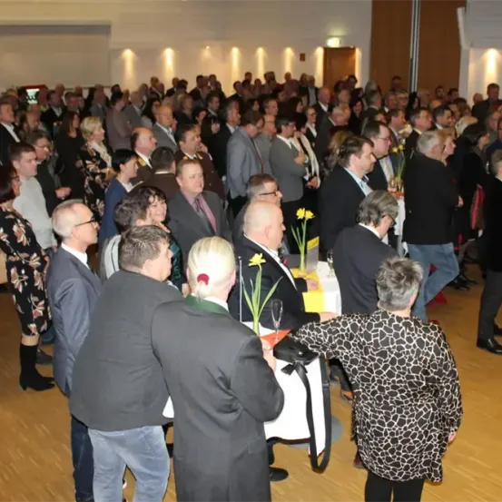 Eine große Gruppe von Menschen ist in einem Raum für eine formelle Veranstaltung versammelt. Viele führen Gespräche, während andere Tische mit gelben Blumen betrachten. Der Raum hat Holzböden und hohe Decken.