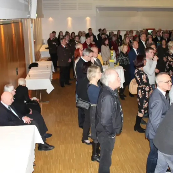 Eine Menschenmenge ist drinnen, wahrscheinlich bei einer formellen Veranstaltung. Sie stehen, sitzen oder unterhalten sich, einige halten Getränke in der Hand. Der Raum hat Holzböden und weiße Wände, mit aufgestellten Tischen und Stühlen.