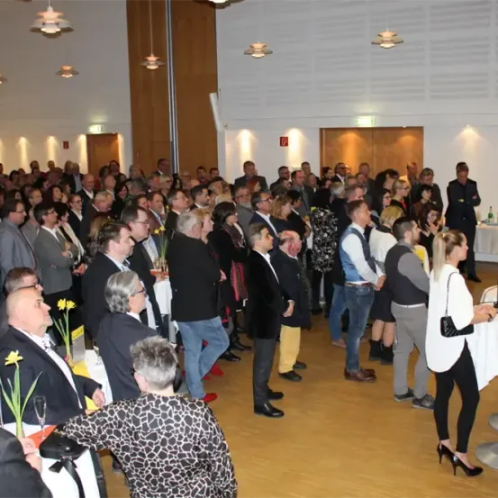 Eine große Gruppe von Menschen versammelt sich drinnen, einige stehen, andere sitzen, mit einem Tisch, auf dem ein Laptop steht. Der Raum ist gut beleuchtet mit mehreren Lampen an der Decke.