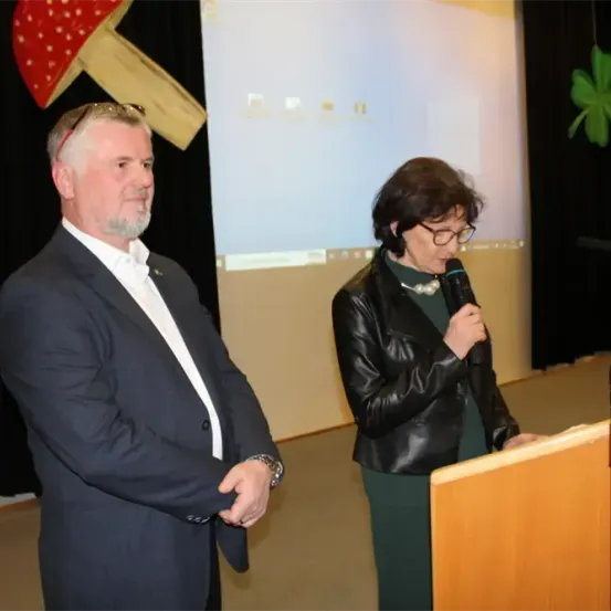 Ein Mann und eine Frau stehen an einem Podium mit einem Mikrofon und einem Bildschirm dahinter. Der Mann trägt einen Anzug und eine Brille auf dem Kopf. Die Frau trägt eine schwarze Lederjacke und spricht in das Mikrofon.