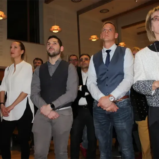 Eine Gruppe von Menschen in formeller Kleidung steht zusammen, möglicherweise bei einer Veranstaltung. Ein Mann mit einer Uhr schaut nach oben, während andere nach vorne blicken. Eine Frau mit Brille steht rechts.