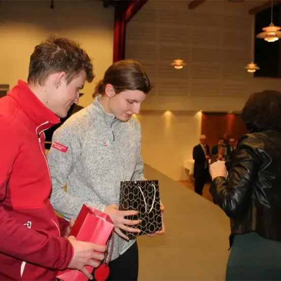 Zwei junge Erwachsene tauschen Geschenke in einem Raum aus, während andere zusehen. Ein Mann in einem roten Jackett hält eine rote Geschenktüte. Die Frau im grauen Sweatshirt hält eine schwarze Geschenktüte.