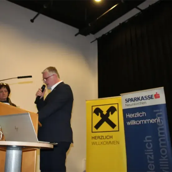 Ein Mann in Anzug steht an einem Podium mit Mikrofon. Ein Laptop liegt auf dem Podium. Hinter ihm steht ein Schild mit 'Herzlich willkommen'. Eine Frau sitzt zu seiner linken Seite. Ein Schild mit 'SPARKASSE Neunkirchen' befindet sich hinter ihnen.