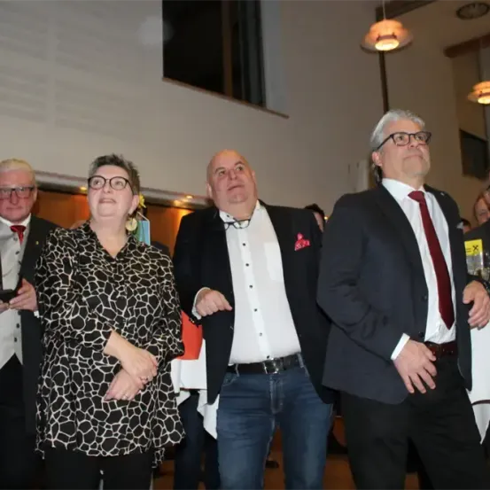 Eine Gruppe formell gekleideter Menschen, darunter eine Frau mit Brille, steht in einem Veranstaltungsort. Sie scheinen auf eine Ankündigung zu warten.