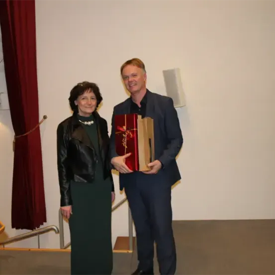 Ein Mann und eine Frau stehen drinnen, lächeln und halten eine Geschenkbox. Die Frau trägt eine Lederjacke und eine Halskette. Der Mann trägt einen Anzug. Eine weiße Wand und rote Vorhänge sind hinter ihnen.