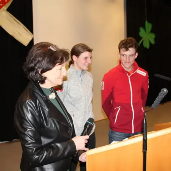 Eine Frau in einer schwarzen Lederjacke spricht in ein Mikrofon, während eine Frau und ein Mann hinter ihr stehen. Der Mann trägt ein rotes Sweatshirt mit einem Logo auf der linken Seite. Dahinter hat eine Wand einen schwarzen Hintergrund mit einem grünen Kleeblattsymbol.