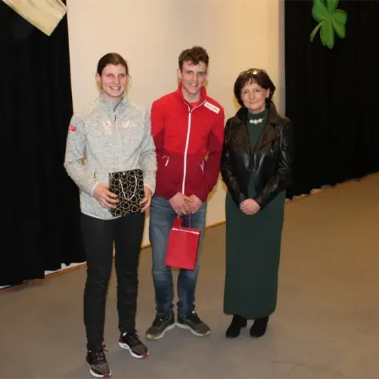 Drei Personen stehen nebeneinander in einem Raum und lächeln für ein Foto. Die Frau auf der linken Seite hält eine schwarze Handtasche. Der Mann in der Mitte hält eine rote Mappe. Die Frau auf der rechten Seite trägt ein grünes Kleid.