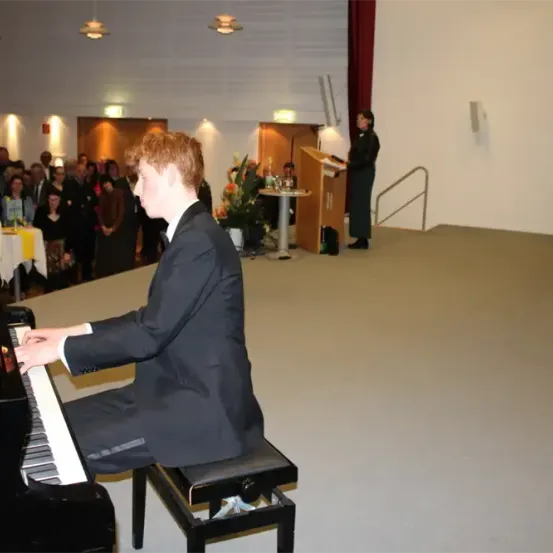 Ein Mann spielt Klavier, während ein Publikum zusieht, und eine Frau spricht von einem Podium aus.