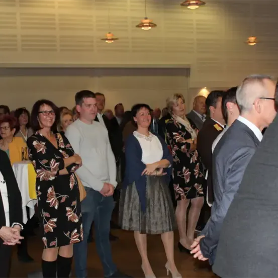 Eine Gruppe von Menschen steht in einem Raum. Einige tragen Brillen, und einige tragen hohe Absätze. Auf der linken Seite steht ein Tisch, und einige Menschen stehen dahinter.