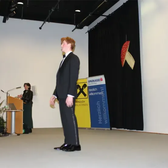 Ein junger Mann in formeller Kleidung steht auf der Bühne mit einem Podium hinter ihm. Eine Frau spricht am Podium. Ein gelber Banner ist im Hintergrund zu sehen.