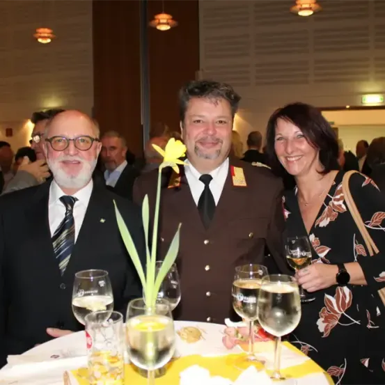 Drei Personen, zwei Männer und eine Frau, lächeln und posieren für ein Foto. Sie sind formell gekleidet, und der Mann in der Mitte hält eine gelbe Blume. Sie stehen vor einem Tisch mit Weingläsern, und es stehen Leute hinter ihnen.