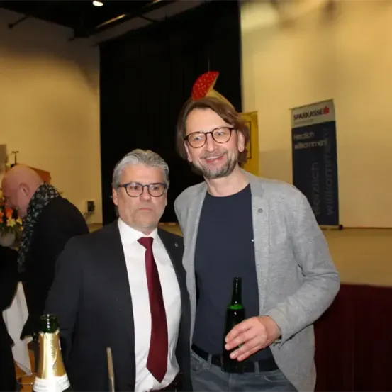 Zwei Männer stehen bei einer Veranstaltung zusammen, einer mit einer Flasche Champagner und der andere mit einem Bier. Sie lächeln, mit einer Bühne und einem Banner hinter ihnen.