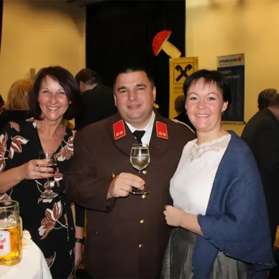 Ein Mann und zwei Frauen lächeln und halten Weingläser. Sie stehen neben einem Tisch mit einem Glas Bier. Dahinter sind mehrere Leute anwesend und ein Schild hängt an der Wand.