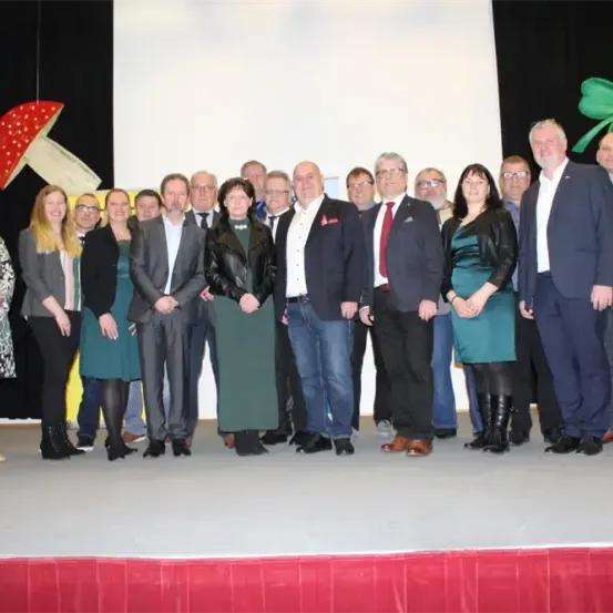 Eine Gruppe von Personen, bestehend aus Männern und Frauen, steht auf einer Bühne, wahrscheinlich bei einer Veranstaltung, und trägt formelle Kleidung. Sie sind in zwei Reihen angeordnet. Hinter ihnen hängen zwei Dekorationen, ein Pilz und ein Kleeblatt.