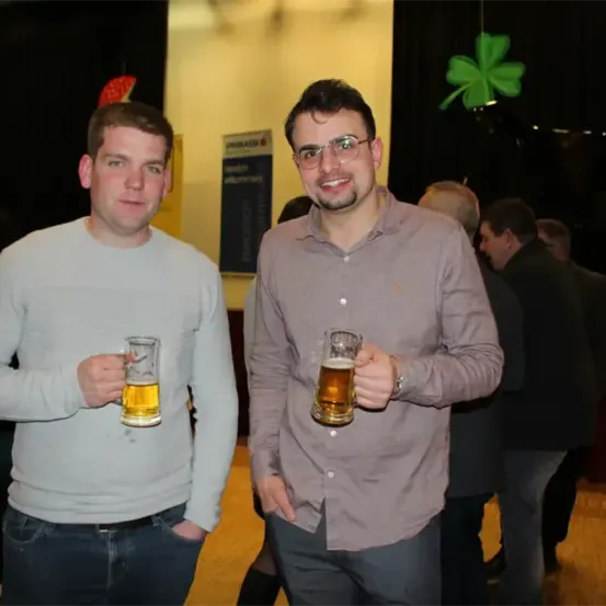 Zwei Männer stehen lächelnd da, jeder hält ein Glas Bier in der Hand. Sie sind lässig gekleidet, einer trägt ein weißes Shirt, der andere ein graues Langarmhemd. Hinter ihnen stehen oder gehen einige Leute.