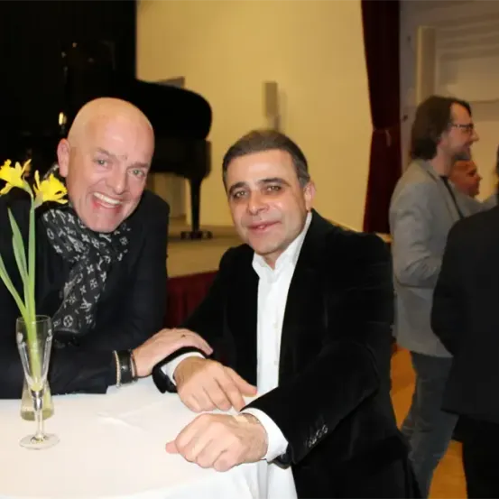 Zwei Männer sitzen an einem Tisch mit einer Blume in einer Vase. Hinter ihnen steht ein Klavier und Menschen stehen und unterhalten sich.