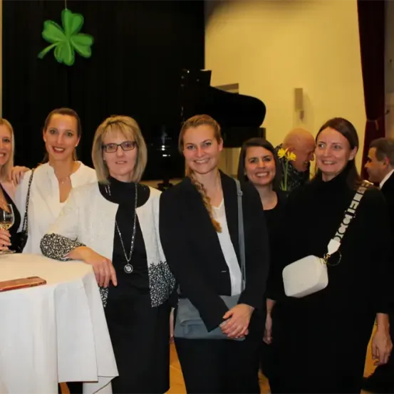 Eine Gruppe von Frauen steht in einem Raum und lächelt für ein Foto. Sie stehen neben einem Tisch mit einem Weinglas und einem schwarzen Klavier im Hintergrund.