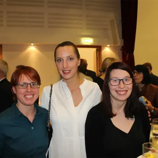 Drei Frauen lächeln und posieren für ein Foto bei einer Veranstaltung. Die Frau in der Mitte trägt eine weiße Bluse. Hinter ihnen stehen andere Leute und unterhalten sich. Der Raum ist schwach beleuchtet, und im Hintergrund befindet sich eine Tür.