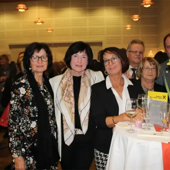 Fünf Frauen stehen mit Weingläsern und einer Blume an einem Tisch, lächeln und posieren für ein Foto in einem Saal.