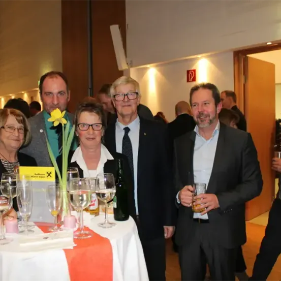 Eine Gruppe Erwachsener in formeller Kleidung, einschließlich Brillen und Krawatten, steht um einen Tisch mit Weinglasern und einer Blume, möglicherweise bei einer Veranstaltung.